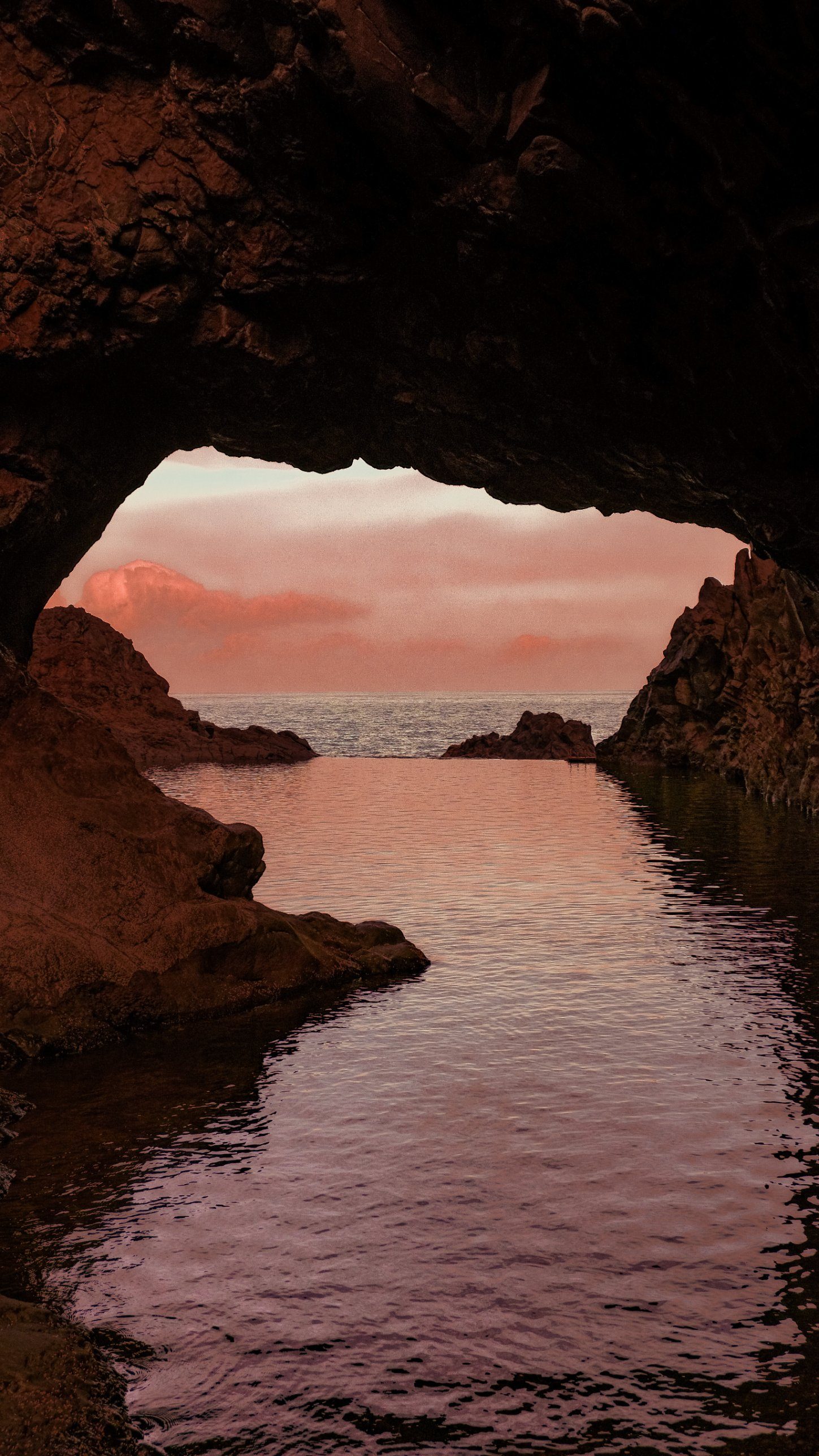 Looking out from inside a rock arch over a still sea pool at sunset, with pink clouds above the horizon