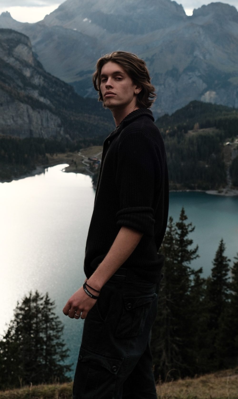 Portrait of Lucas Hunt in a dark knit cardigan, standing on a mountainside above an alpine lake and glancing at the camera
