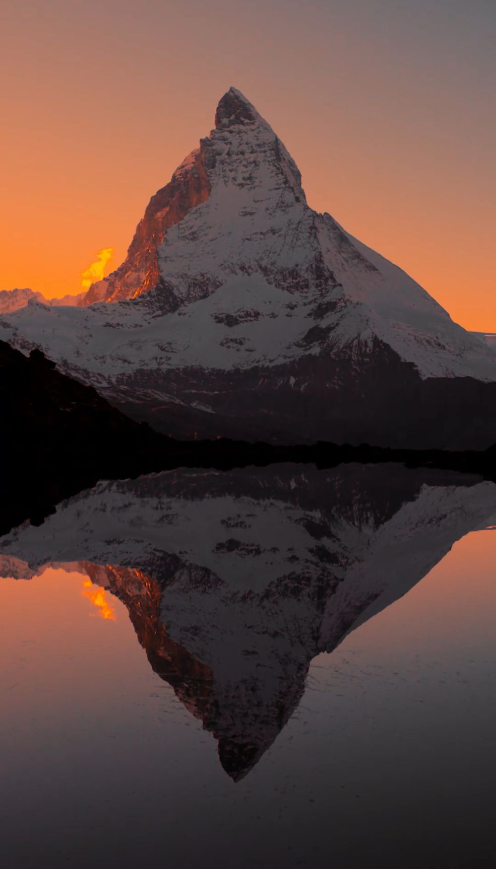 A snow-capped pointed peak glowing orange at sunset, mirrored in a still lake